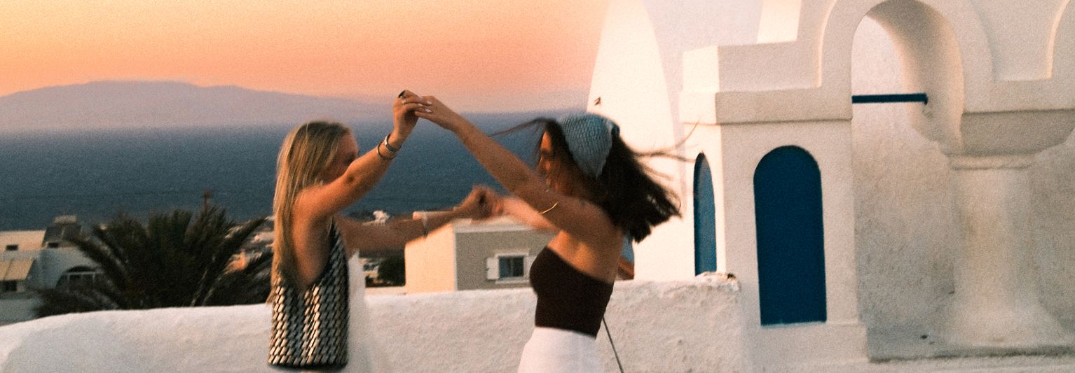 Two people joyfully dancing on a white rooftop in a coastal town at sunset, with the sea and buildings in the background.