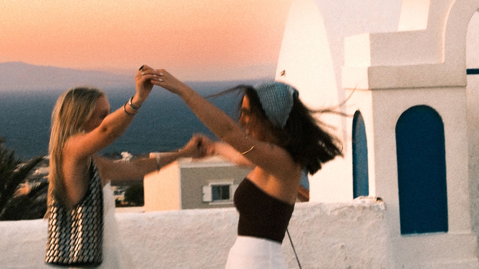 Two people joyfully dancing on a white rooftop in a coastal town at sunset, with the sea and buildings in the background.