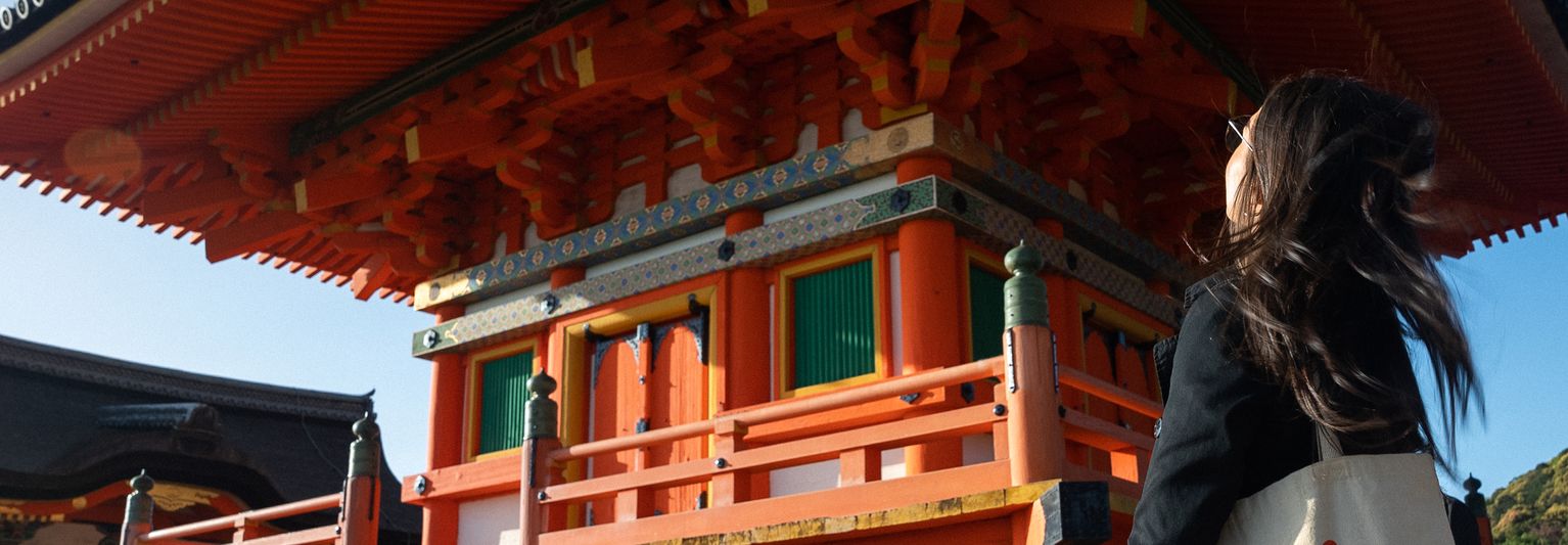A person stands gazing at a traditional Japanese pagoda, with intricate wooden architecture and vibrant red and orange colors, under a clear blue sky.