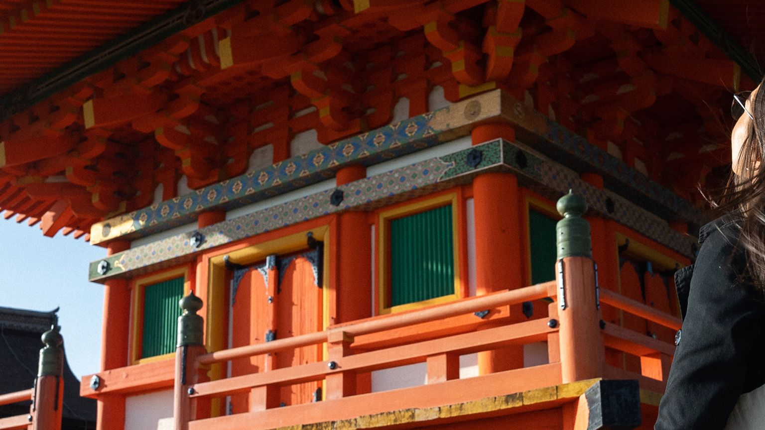 A person stands gazing at a traditional Japanese pagoda, with intricate wooden architecture and vibrant red and orange colors, under a clear blue sky.