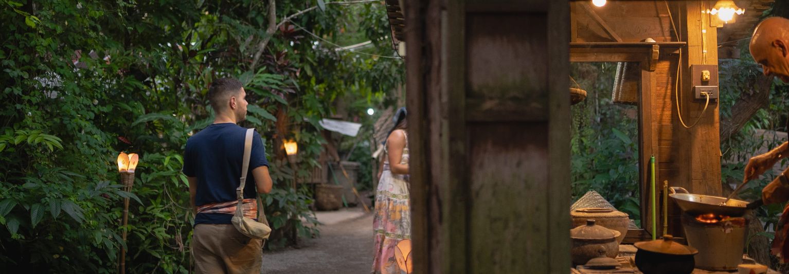 A man walking down a path through lush greenery in Thailand past a man cooking in an open-air stall