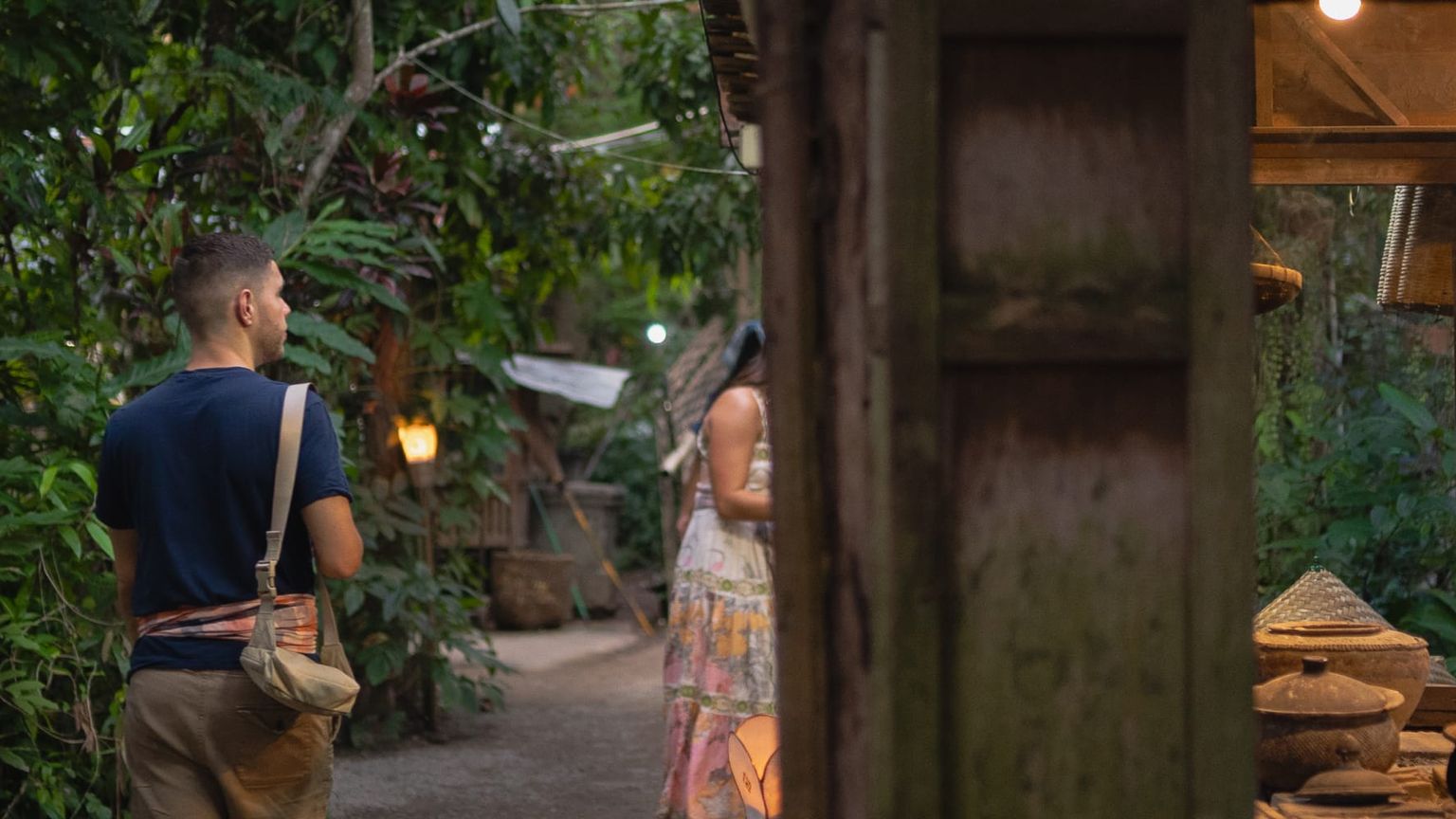 A man walking down a path through lush greenery in Thailand past a man cooking in an open-air stall