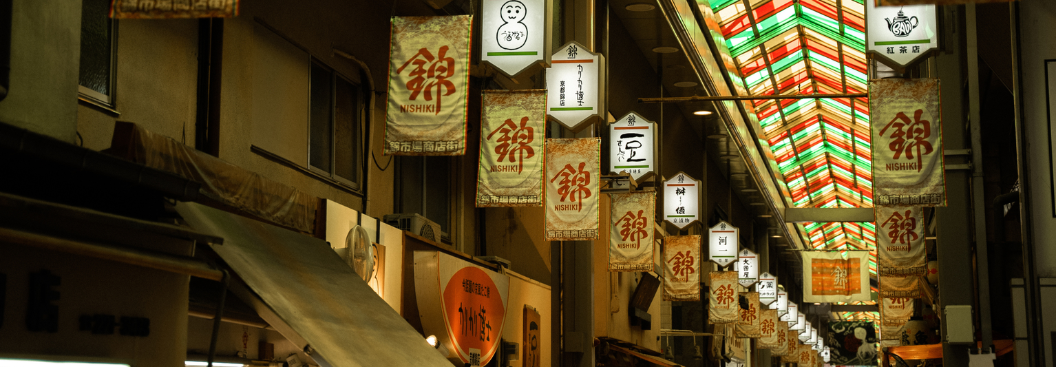 A vibrant indoor market with colorful lanterns and signs hanging from the ceiling, featuring a variety of symbols and Japanese characters.
