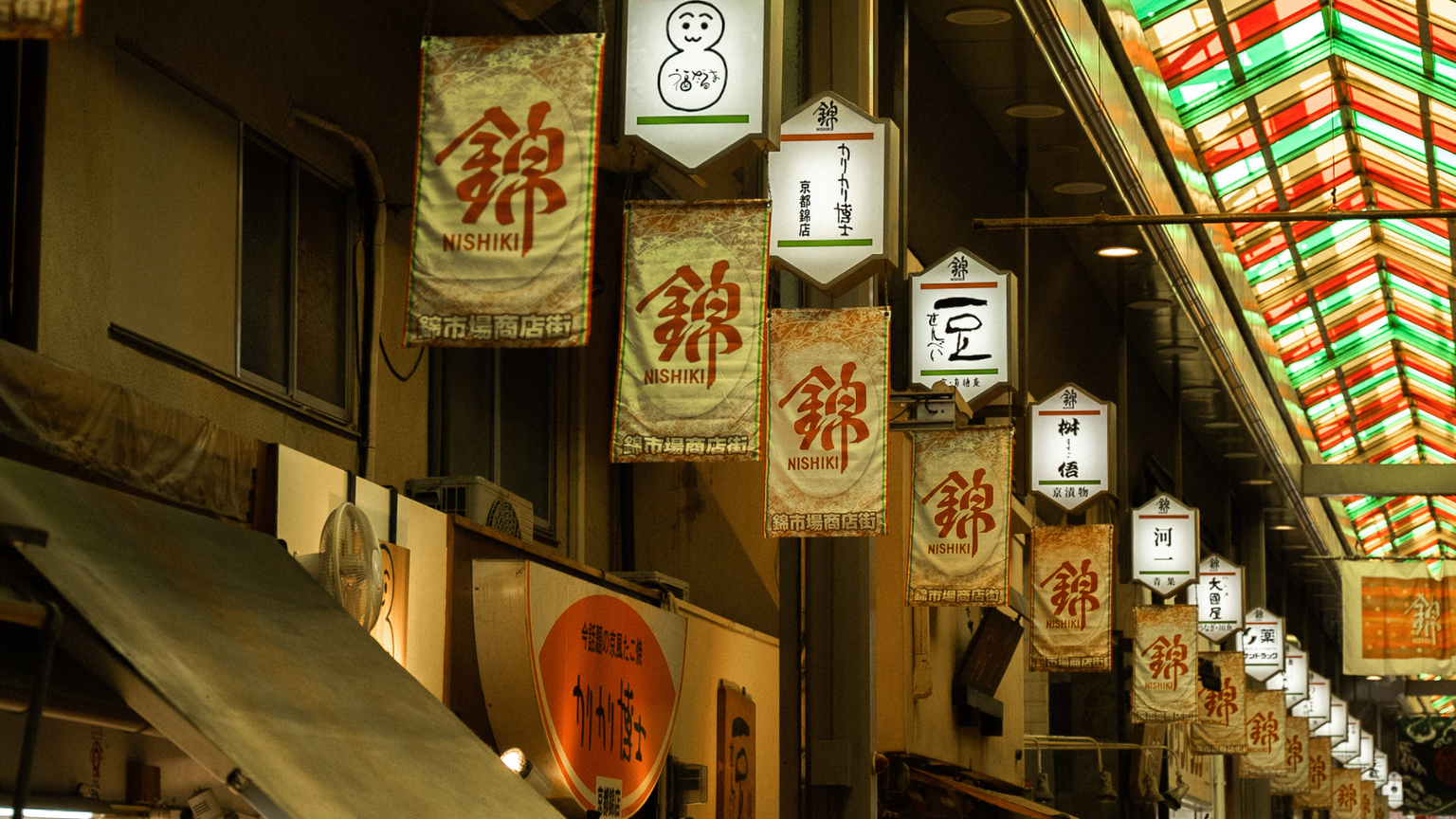 A vibrant indoor market with colorful lanterns and signs hanging from the ceiling, featuring a variety of symbols and Japanese characters.