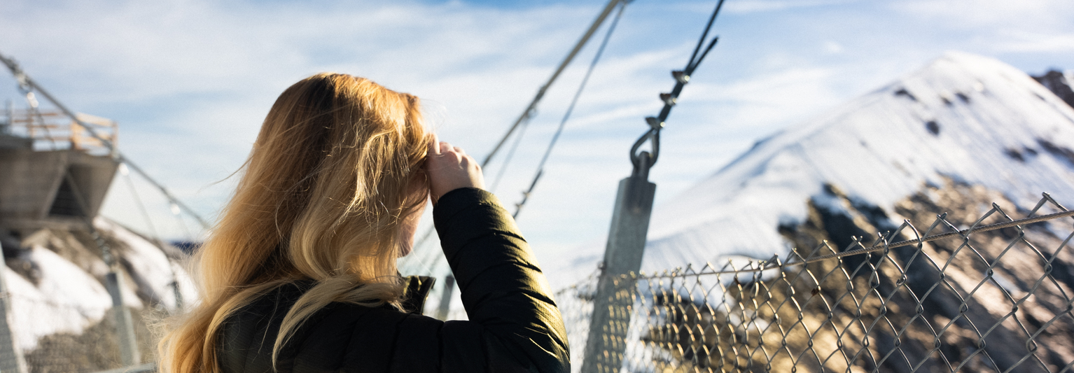 Woman with long blonde hair stands on a snowy mountain bridge, looking at the view, with a fence and cables in the foreground.