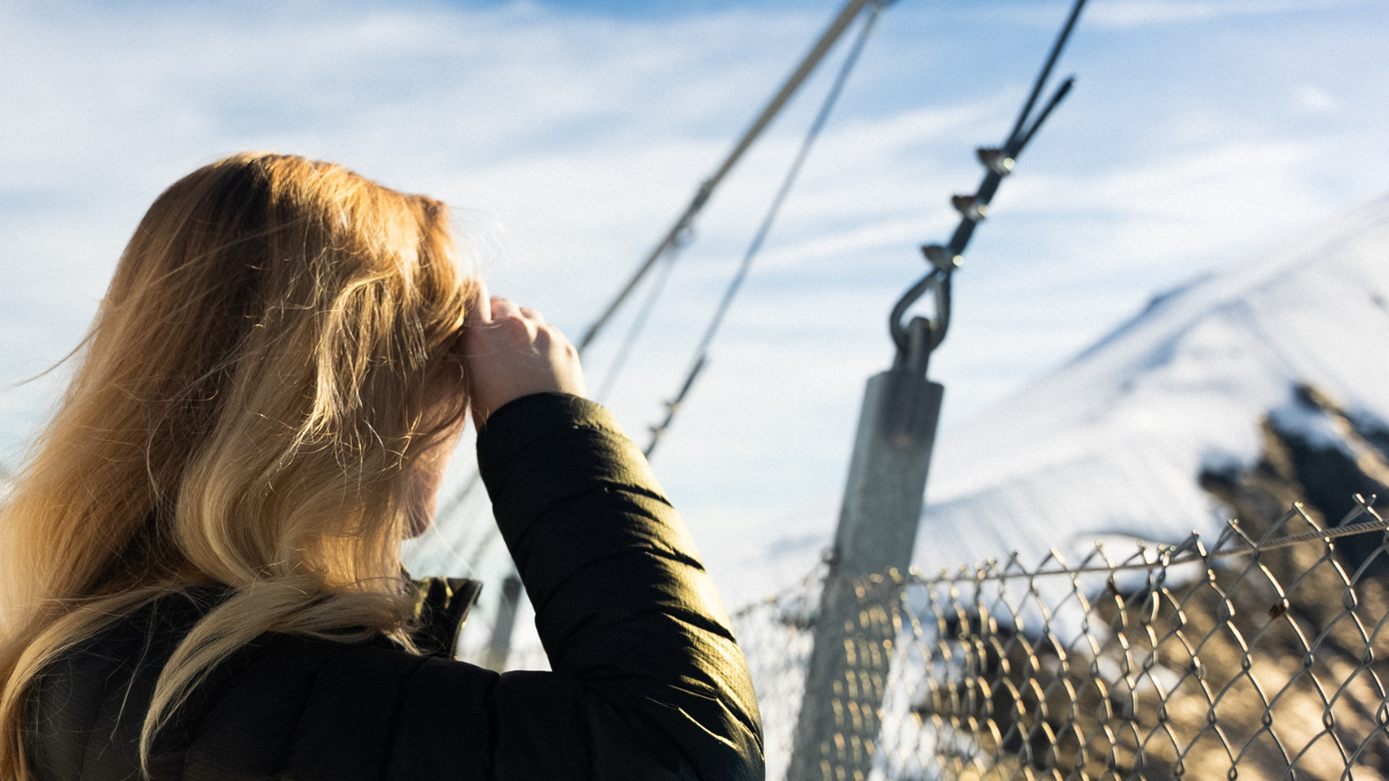 Woman with long blonde hair stands on a snowy mountain bridge, looking at the view, with a fence and cables in the foreground.