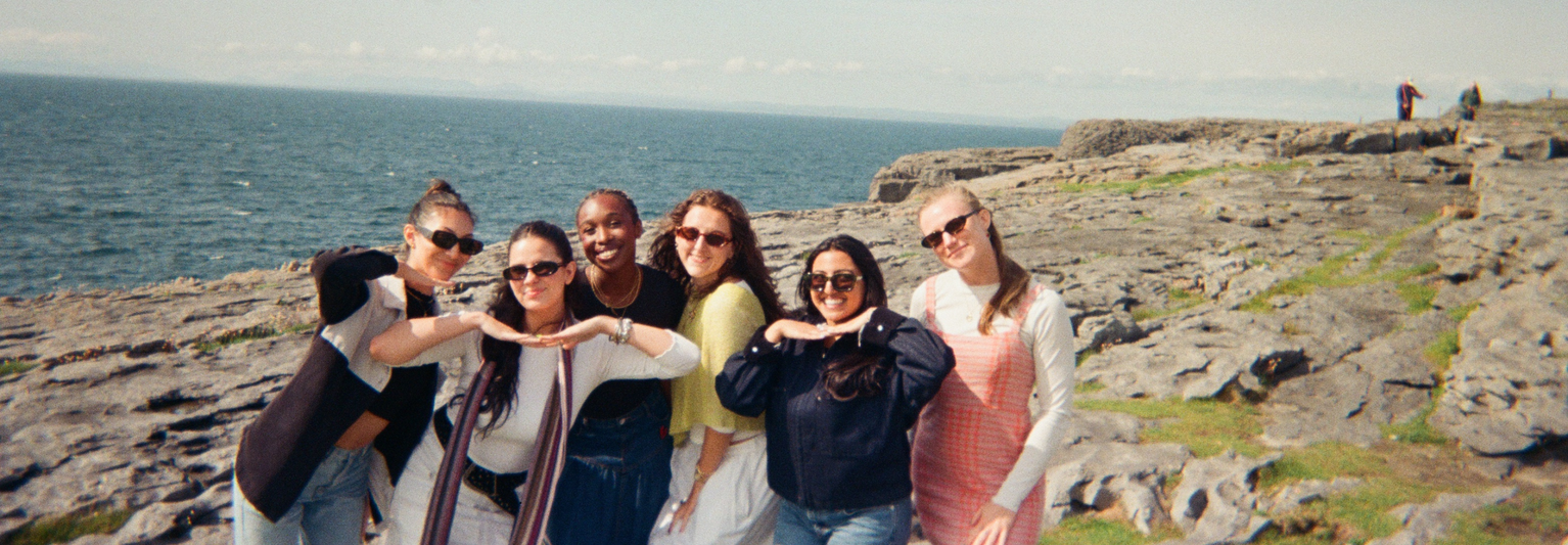 Six friends pose playfully on a rocky seaside cliff with the ocean in the background under a clear sky.