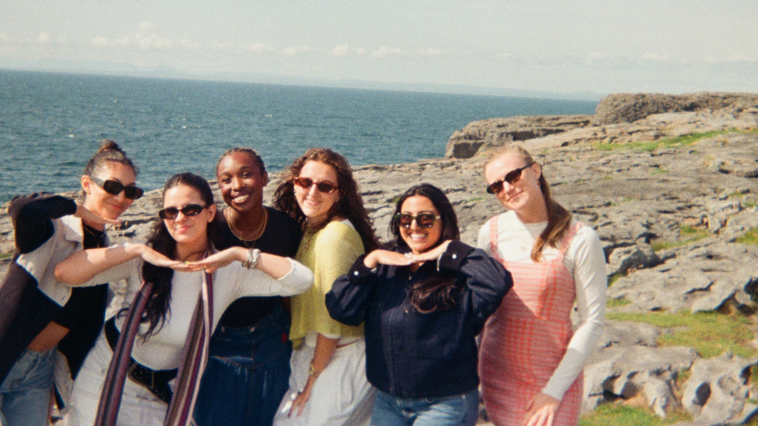 Six friends pose playfully on a rocky seaside cliff with the ocean in the background under a clear sky.