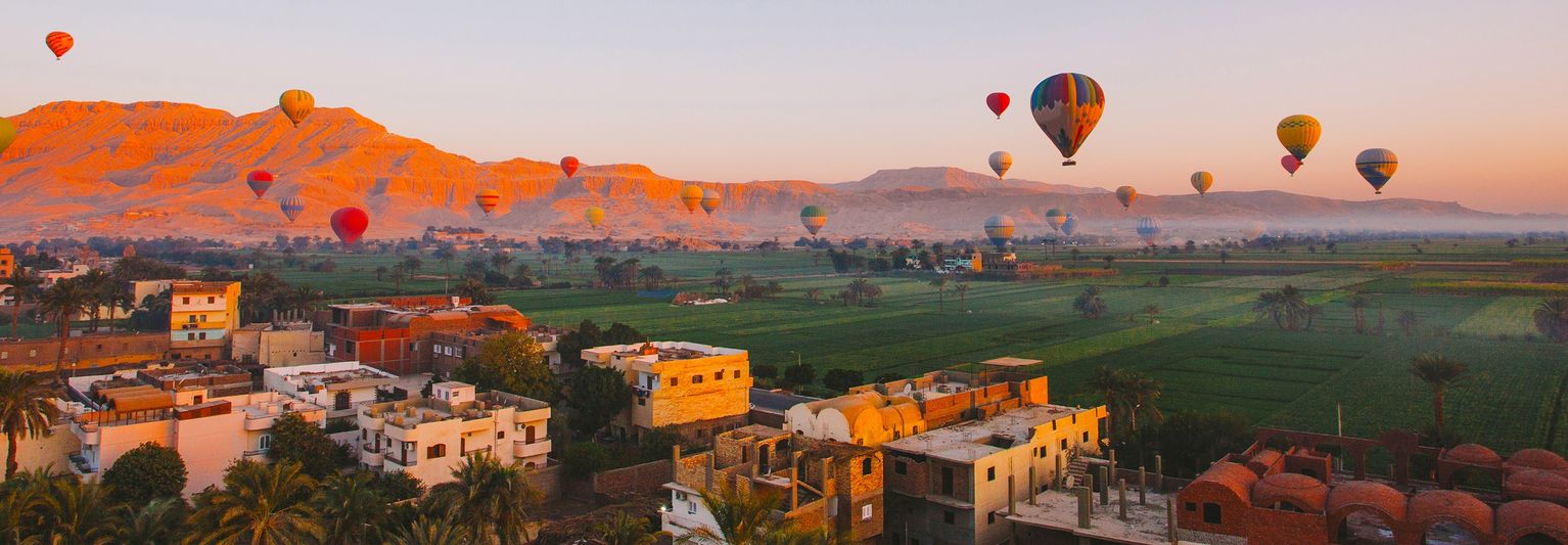 Many hot air balloons floating over buildings, a green field, and mountains at sunset in Luxor, Egypt