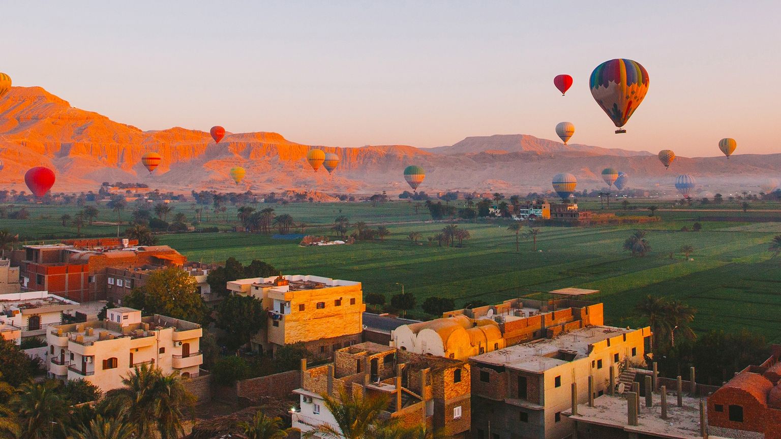 Many hot air balloons floating over buildings, a green field, and mountains at sunset in Luxor, Egypt