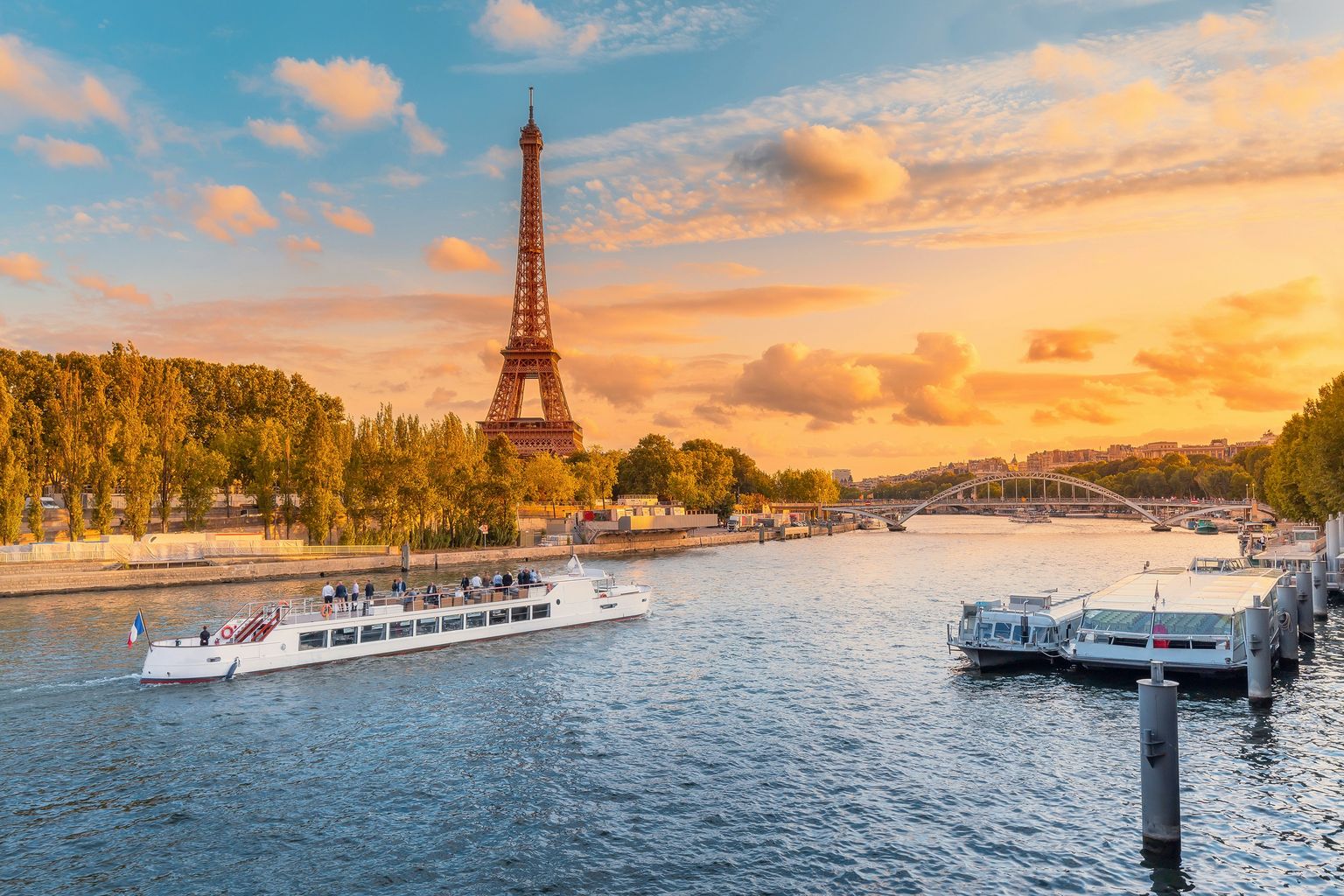 A river cruise boat sails on the Seine at sunset, with the Eiffel Tower and the Parisian skyline in the background.
