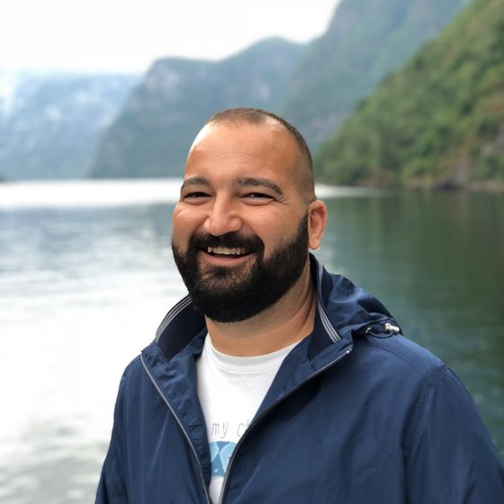 A man in a blue jacket smiles near a calm lake, surrounded by tree-covered hills under an overcast sky.