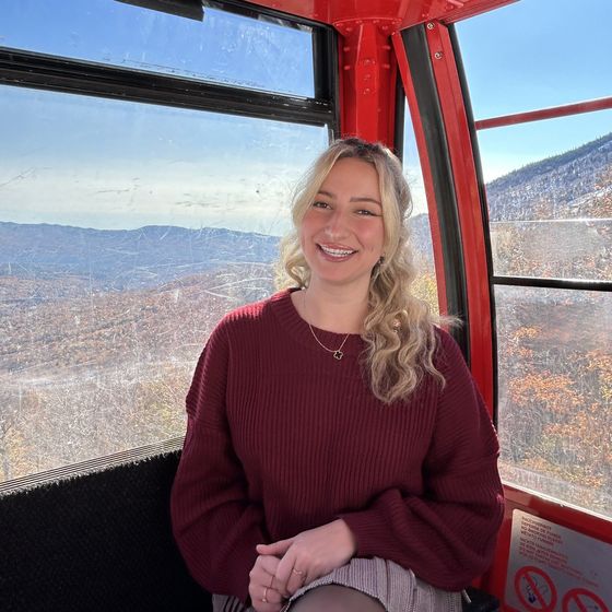 A woman in a red sweater smiling inside a red cable car with a scenic mountain and autumn landscape visible through the window.