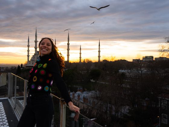 A woman smiles on a balcony with a mosque in the background at sunset, under a cloudy sky with flying birds.