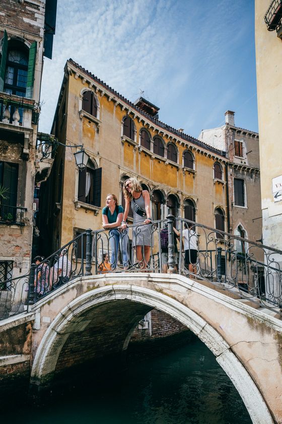Three people stand on a stone arch bridge over a canal, surrounded by historic buildings on a sunny day.