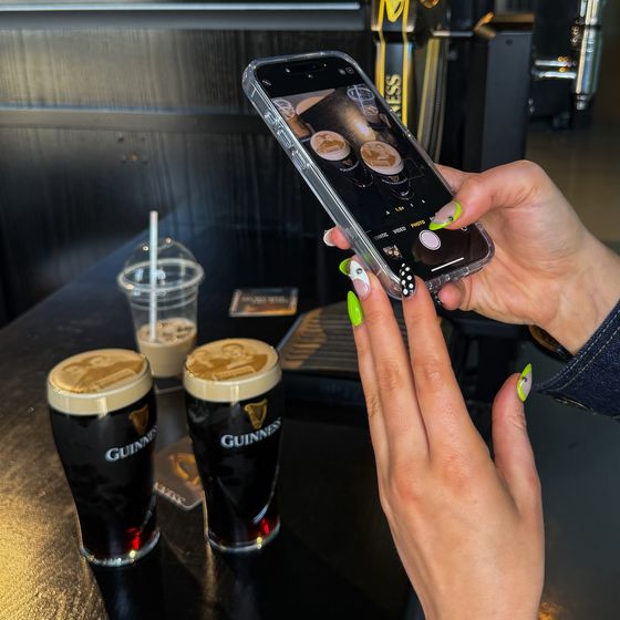 A person with green nails takes a photo of two pints of Guinness on a table, capturing them with a smartphone.