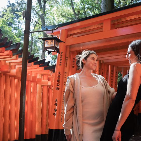 Two women smiling at each other while standing beneath red torii gates in a sunlit path at Fushimi Inari Shrine, Kyoto.