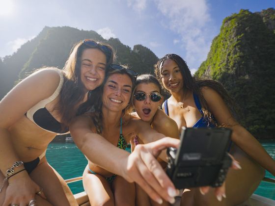 Four women in swimsuits take a selfie on a boat with turquoise water and rocky, lush green cliffs in the background under a clear blue sky.