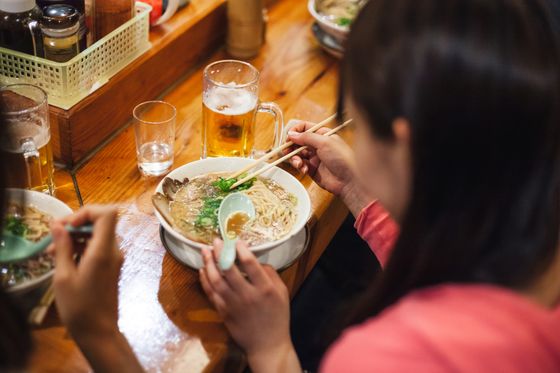 A person in a red shirt eats ramen using chopsticks at a wooden table, with a glass of beer nearby.