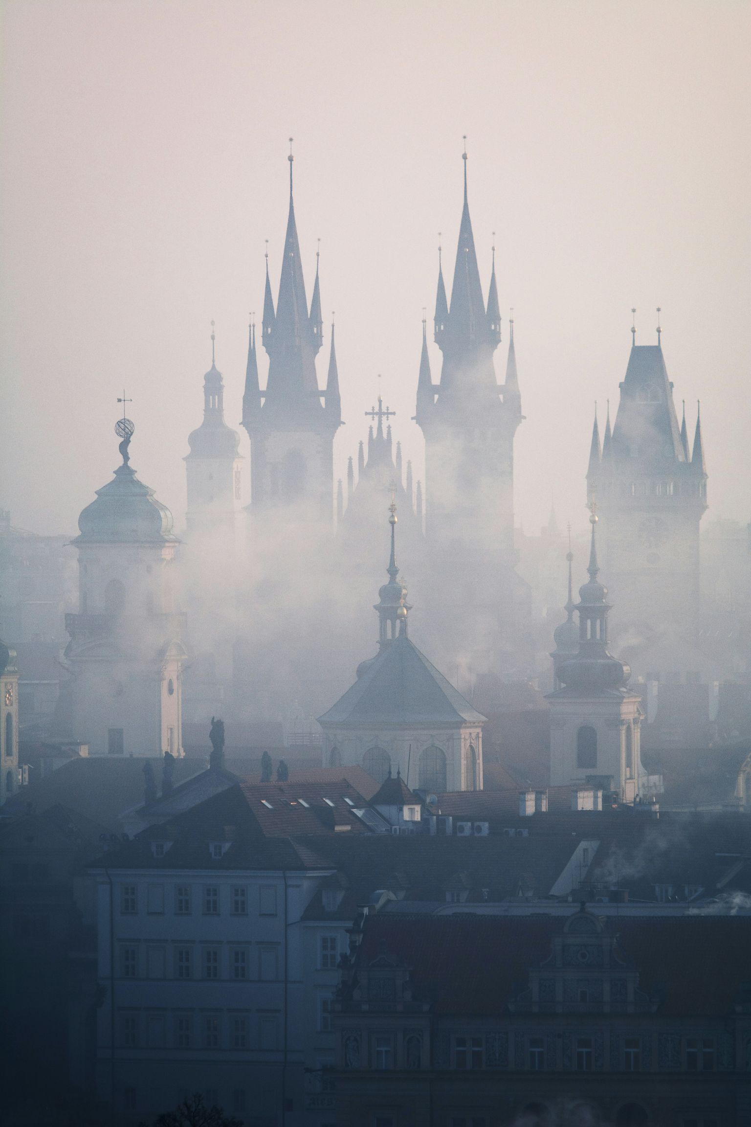Silhouetted Gothic spires and domes rise through thick mist over an old European cityscape at dawn.