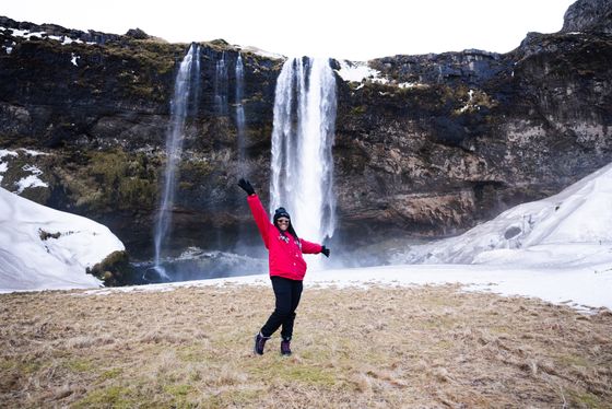 Person in red jacket joyfully poses with arms up in front of a tall waterfall cascading down a rocky cliff, surrounded by snowy and grassy landscape.