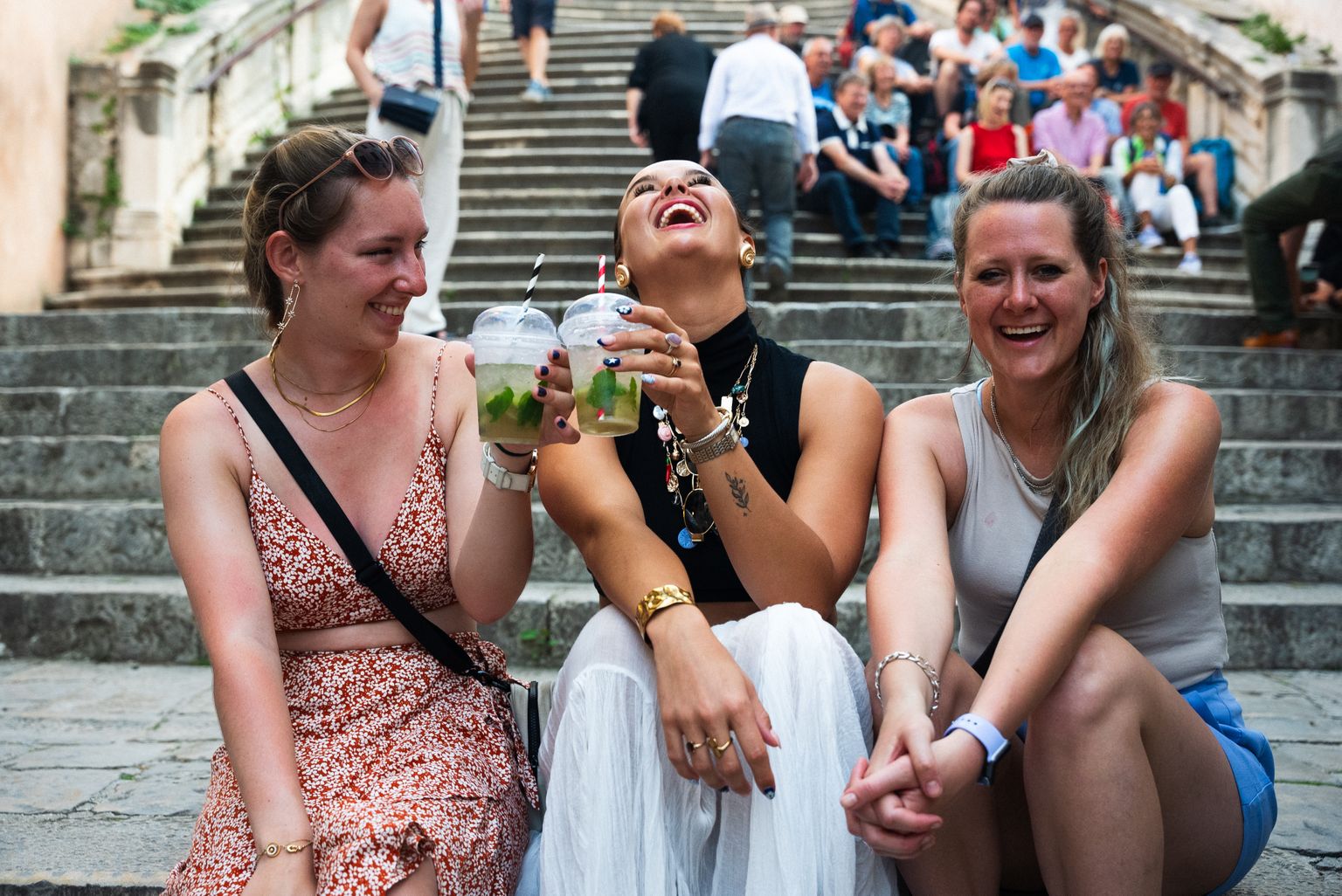 Three women sitting on stone steps, laughing and holding drinks, with people in the background.