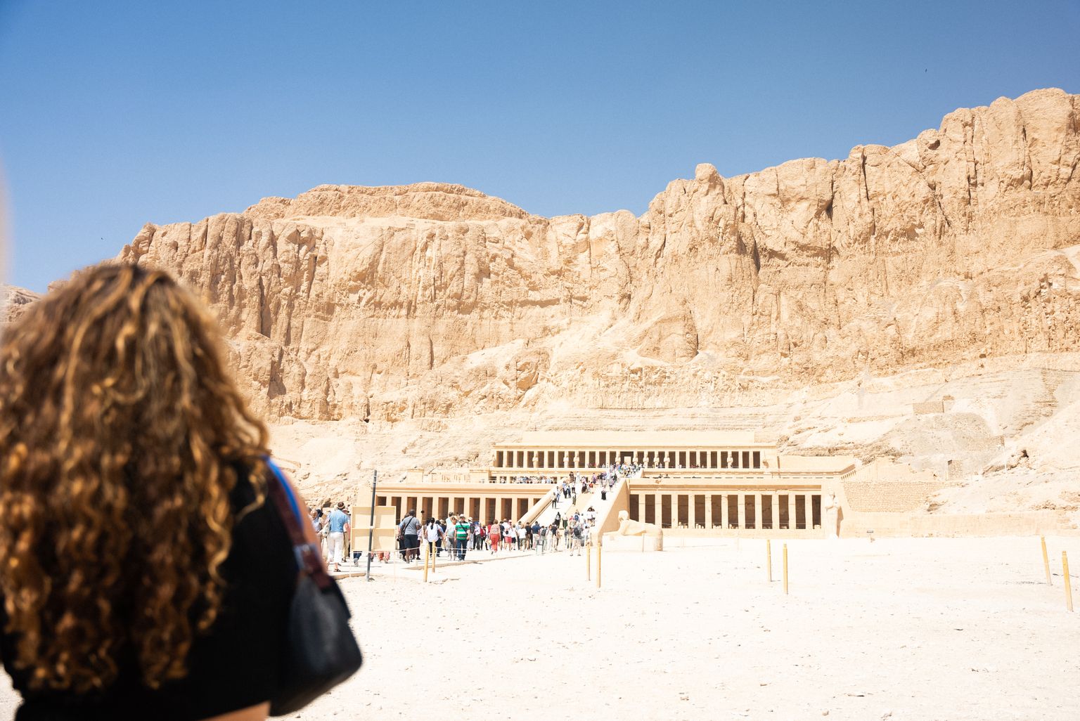 A woman with curly hair looks at the ancient Temple of Hatshepsut, nestled against a rocky cliff under a clear blue sky, with tourists approaching.