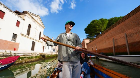 Person rowing a boat through a narrow canal, with a historic building and smiling passengers, under a clear blue sky.