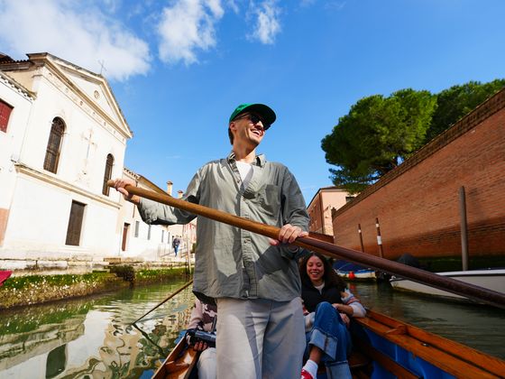 Person rowing a boat through a narrow canal, with a historic building and smiling passengers, under a clear blue sky.