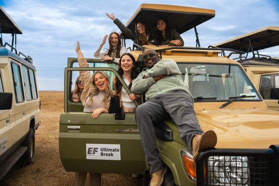 A group of smiling people pose enthusiastically by a safari vehicle in a grassy field, with blue skies above.