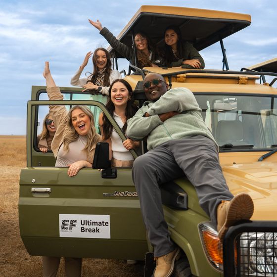 A group of smiling people pose enthusiastically by a safari vehicle in a grassy field, with blue skies above.