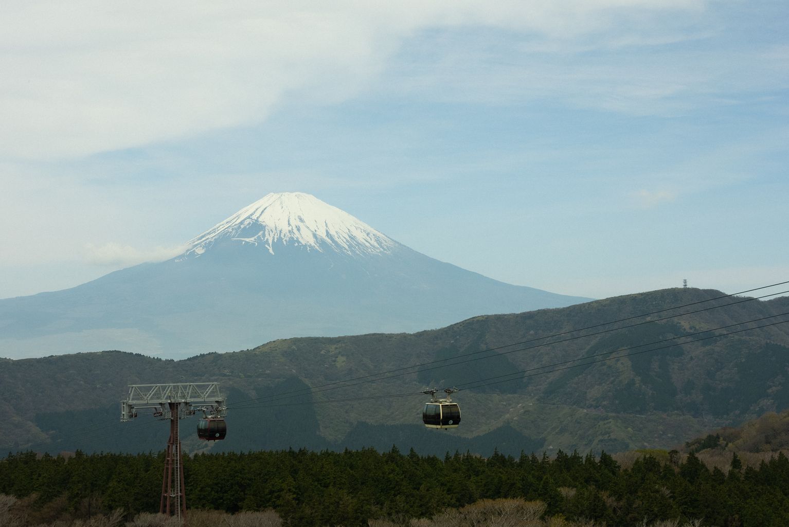 Snow-capped Mount Fuji with cable cars in the foreground, set against a clear sky and lush green landscape.