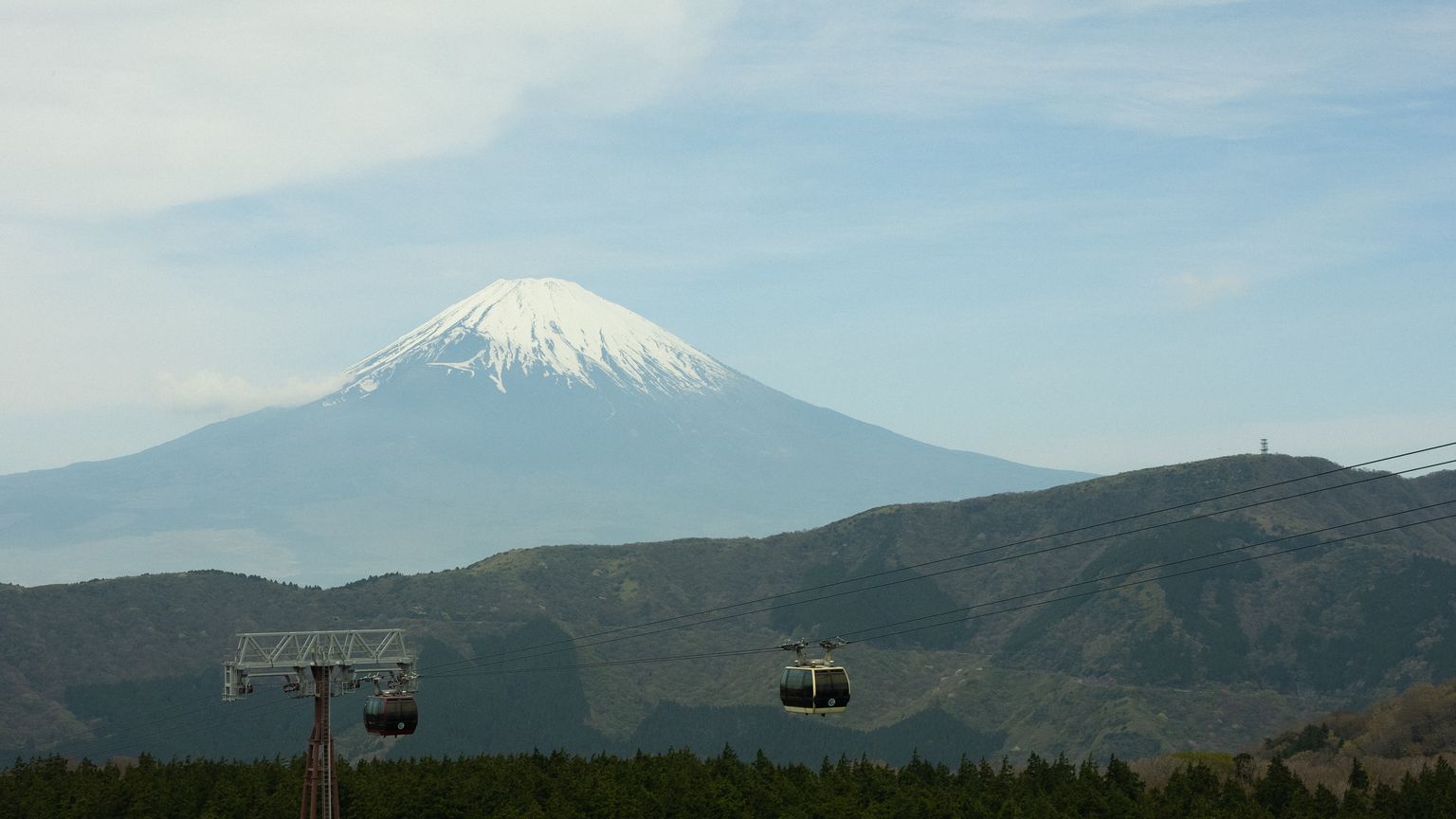 Snow-capped Mount Fuji with cable cars in the foreground, set against a clear sky and lush green landscape.