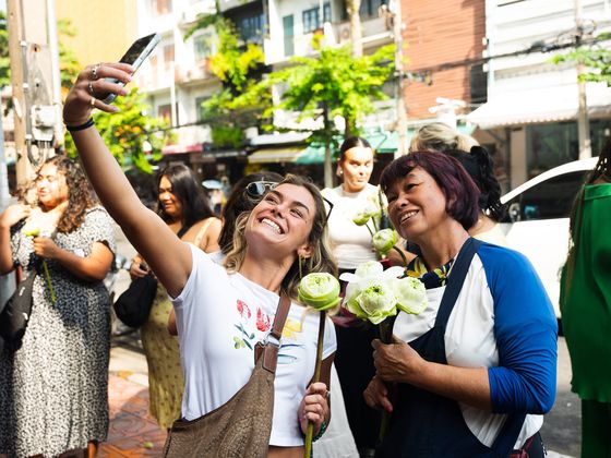 Two people smiling and taking a selfie with lotus flowers on a lively street, surrounded by others.