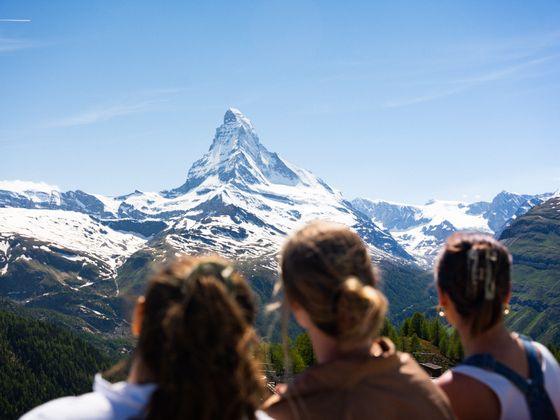 Three people look at the snow-capped Matterhorn under a clear blue sky, surrounded by green and snowy mountain peaks.