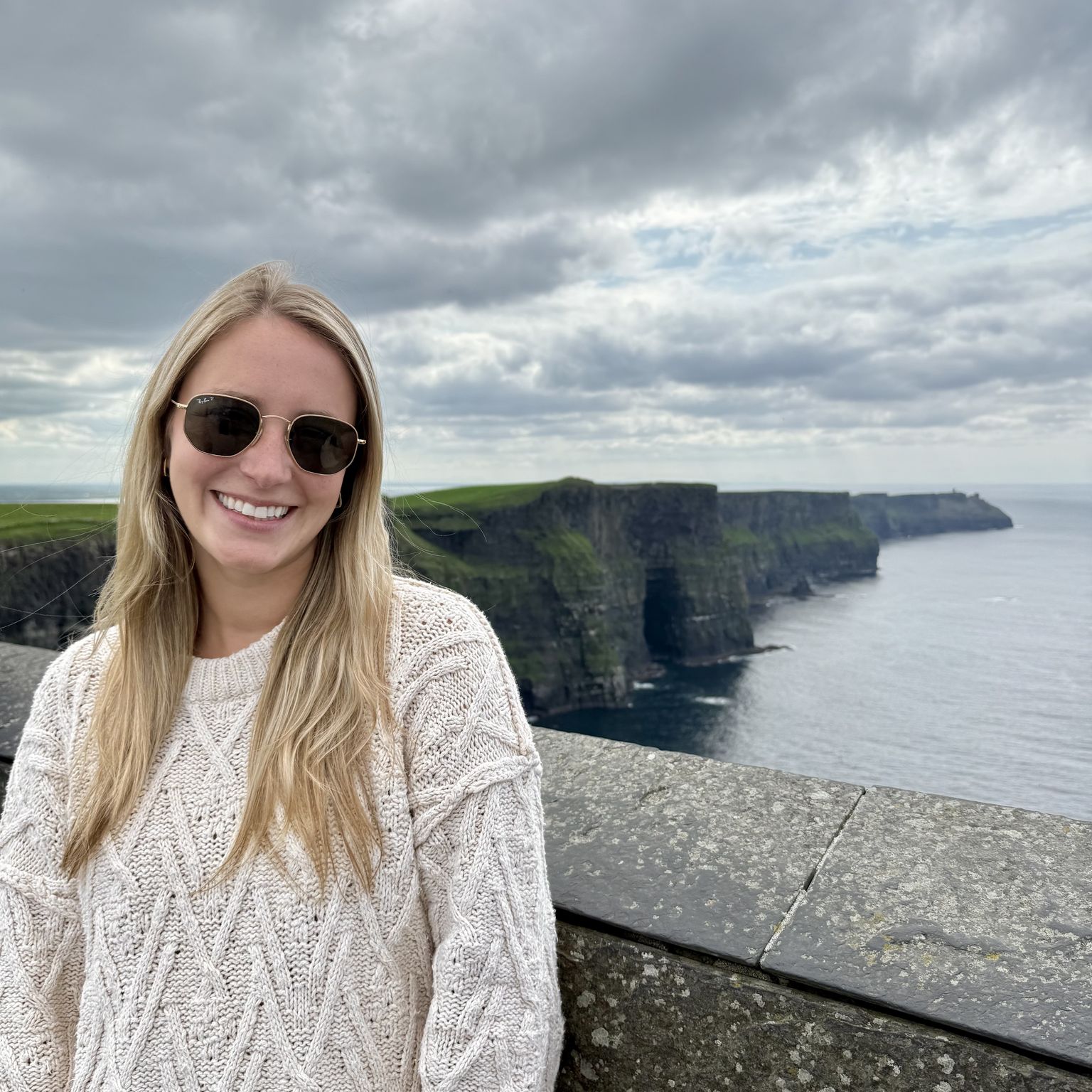 A woman in sunglasses smiling and posing in front of the Cliffs of Moher in Ireland