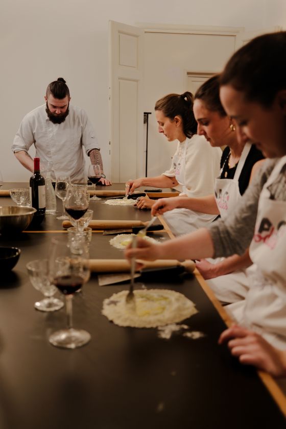A chef guides four people making pasta at a table, each with bowls and a glass of red wine, in a cooking class.