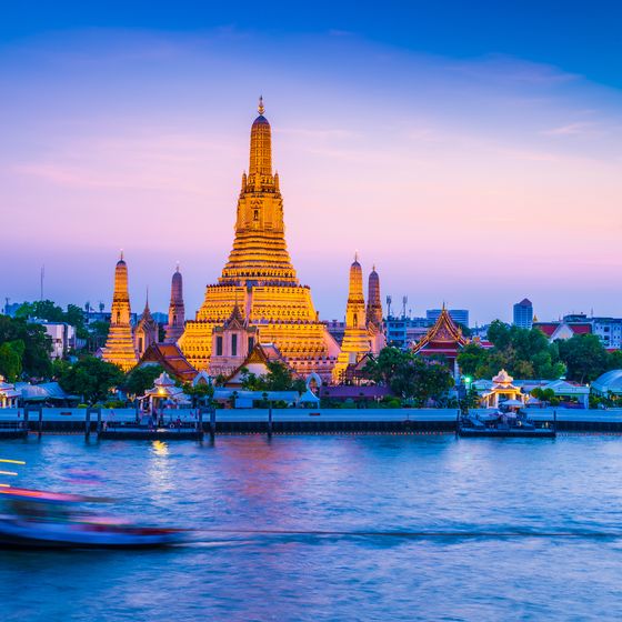 Wat Arun temple illuminated at dusk, with a blurred boat on the Chao Phraya River in the foreground and a cityscape in the background.