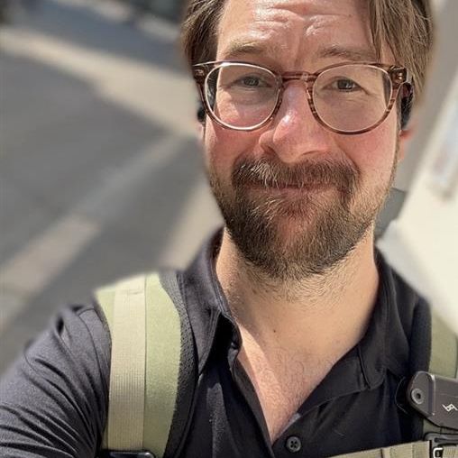 Man with glasses and a beard smiling, wearing a black shirt and backpack, standing outdoors on a sunny day.
