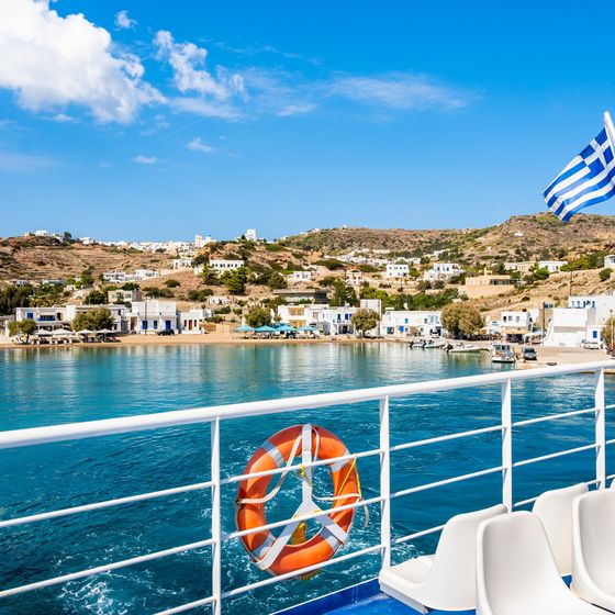 View from a ferry featuring the Greek coastline, blue water, a Greek flag, and nearby village buildings under a clear blue sky.