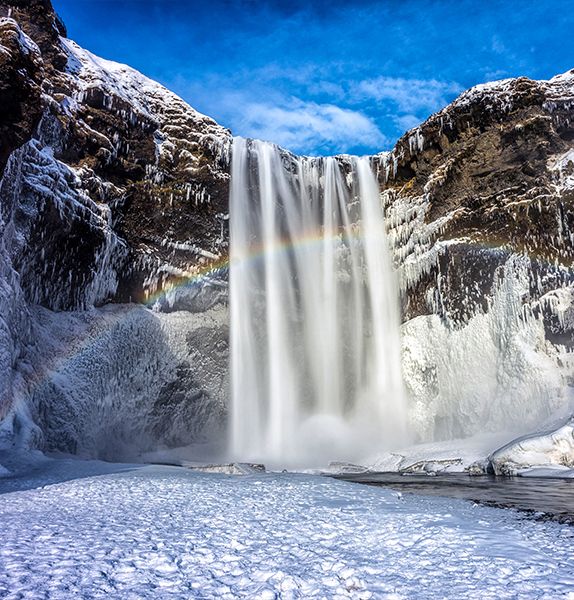 A majestic waterfall cascades over icy cliffs, with a vivid rainbow arching through the mist against a bright blue sky.