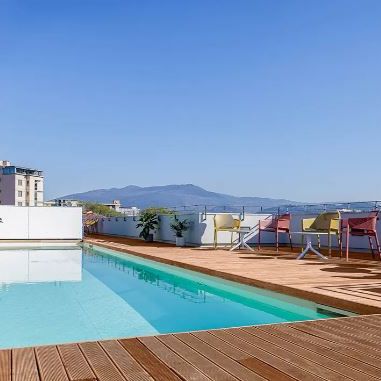 Rooftop pool with wooden deck, tables, and chairs, overlooking a cityscape and mountains under a clear blue sky.