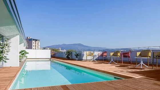 Rooftop pool with wooden deck, tables, and chairs, overlooking a cityscape and mountains under a clear blue sky.