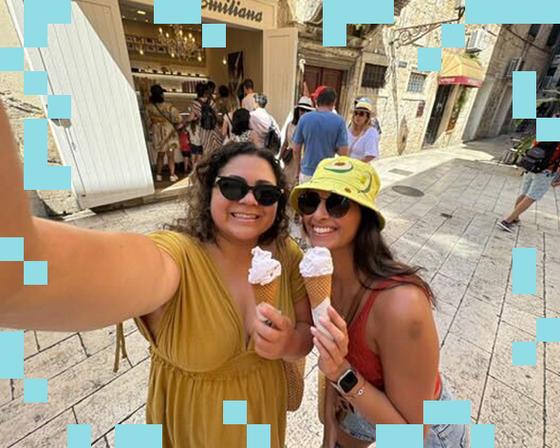 Two women smiling with ice cream cones outside a shop on a sunny day, surrounded by people and historical buildings.