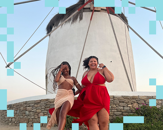 Two people in flowing dresses happily pose in front of a white windmill against a clear sky.