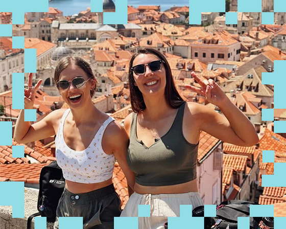 Two women smiling and posing with peace signs against a backdrop of terracotta rooftops in a sunny coastal city.