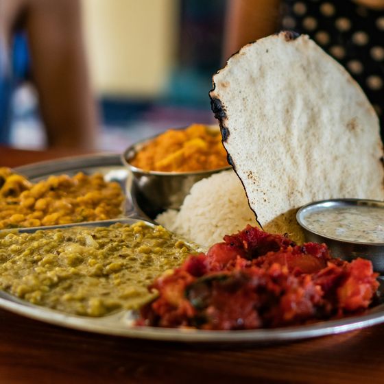 A stainless steel plate with rice, chapati, lentils, chickpeas, potatoes, and a small bowl of raita on a wooden table.