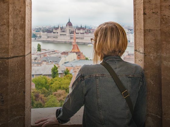 Person in denim jacket looking at a cityscape through a stone archway, with a river and a historic building in the distance.