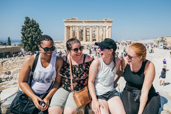 Four women laughing and sitting together at the Acropolis in Athens, with the Parthenon and a crowd in the background under a clear blue sky.