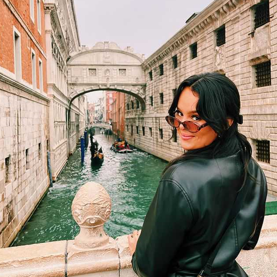 Woman in sunglasses and a black jacket stands by a canal in Venice, with gondolas and the Bridge of Sighs in the background.